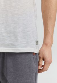 Tricou din bumbac gri deschis cu un finisaj texturat, având o etichetă mică bleumarin și albă la tiv și asociat cu pantaloni de trening gri închis.