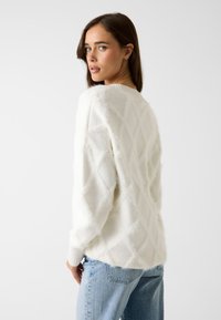 Maglione bianco e soffice con motivo a diamante e polsini a coste, abbinato a jeans azzurri. La texture sembra morbida e accogliente.