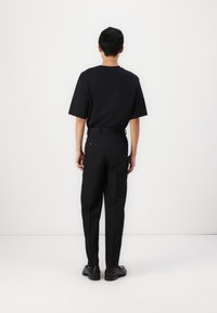 Chemise noire à manches courtes et pantalon noir, vus de l'arrière. Tissu lisse, coupe ajustée et boutons contrastants sur les poches arrière.