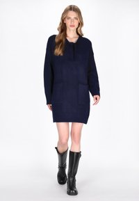 Rochie tricotată bleumarin cu glugă, două buzunare frontale, tiv și manșete cu model canalé. Asortată cu cizme negre până la genunchi.