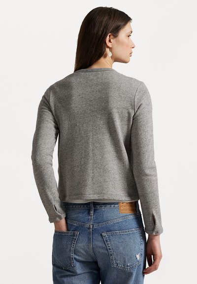 Pullover grigio a maniche lunghe con un knit texturizzato. Design cropped con scollatura tonda e leggere fessure ai polsini delle maniche, indossato con jeans blu.