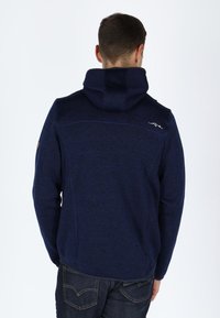 Felpa zip con cappuccio di colore blu navy, con finitura testurizzata, cappuccio e un piccolo logo bianco nella parte superiore della schiena, abbinata a jeans scuri.
