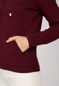 Hoodie bordeaux avec une poche à l'avant, des poignets côtelés et un tissu texturé. Comprend un cordon de serrage avec des embouts métalliques. Coupe simple et décontractée.