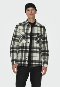 Camisa de franela a cuadros en blanco y negro con dos bolsillos en el pecho, cierre de botones en la parte delantera y una camiseta de manga larga negra debajo. Material de algodón.