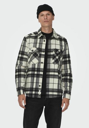 Camisa de franela a cuadros en blanco y negro con dos bolsillos en el pecho, cierre de botones en la parte delantera y una camiseta de manga larga negra debajo. Material de algodón.