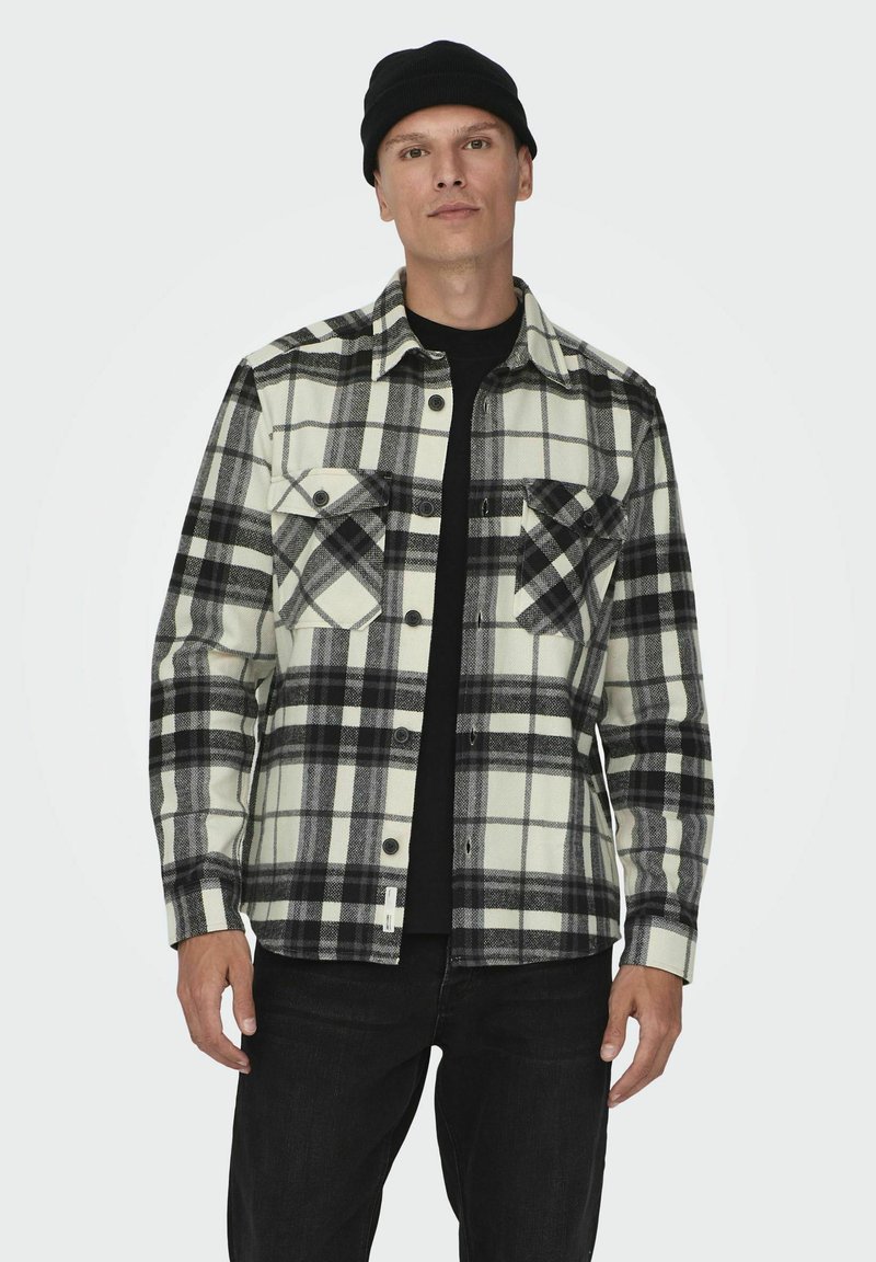 Camisa de franela a cuadros en blanco y negro con dos bolsillos en el pecho, cierre de botones en la parte delantera y una camiseta de manga larga negra debajo. Material de algodón.