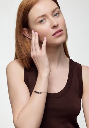 Czarna bransoletka z koralików na gołej nadgarstku, wykonana z gładkiego materiału, z czterema równomiernie rozmieszczonymi prostokątnymi koralikami na delikatnym łańcuszku.