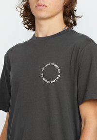 T-shirt en coton gris foncé à manches courtes. Présente un logo circulaire blanc avec du texte à l'avant. Coupe décontractée, encolure simple et texture douce.