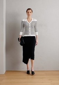 Un cardigan rayé noir et blanc sur une jupe noire, tenant un sac noir texturé. Des chaussures sombres et simples complètent la tenue. Design minimaliste.