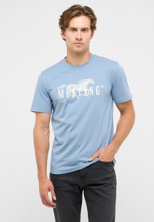 Young man with light brown hair wearing light blue Mustang t-shirt and black pants, standing with one hand in pocket on a plain background.