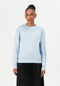 Light blue sweatshirt with ribbed cuffs and hem, featuring embroidered "Tommy Hilfiger" logo on the chest, paired with a black pleated skirt.
