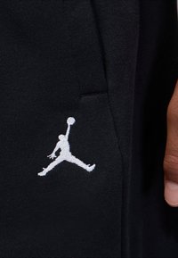 Tissu noir avec un logo Jumpman blanc brodé, représentant une silhouette en train de sauter en tenant un ballon de basket, près du bord d'une poche.