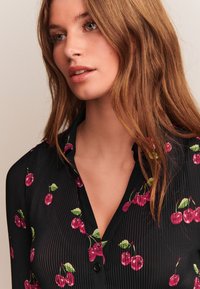 Chemise noire à manches longues avec une texture côtelée verticale, ornée d'un motif de cerise en rouge et vert. Comprend un col à revers et des boutons.