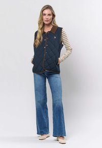 Marineblå quiltet vest med brun kordfløyelskrage og kontrasterende piping, kombinert med stripete langermet skjorte og vide jeans.