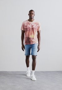 Camiseta de manga corta rosa con gráfico y texto "Bondi", combinada con pantalones cortos de denim azul claro y zapatillas blancas. Textura suave, diseño casual.
