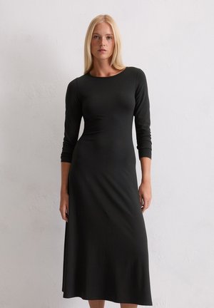 Vrouw met lang blond haar in een aansluitende zwarte midi-jurk met lange mouwen, die tegen een witte tekstuurmuur staat.