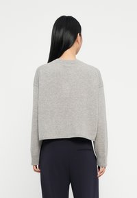 Frau mit langen schwarzen Haaren, die einen grauen kurzen Pullover und dunkle Hose trägt, steht mit dem Rücken gegen einen schlichten weißen Hintergrund.