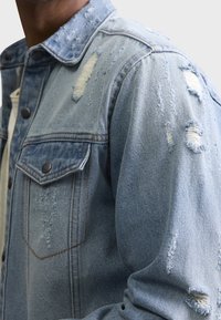 Chaqueta de mezclilla azul clara con detalles desgastados, que cuenta con un cuello clásico, cierre de botones, bolsillos en el pecho y acentos deshilachados en las mangas.