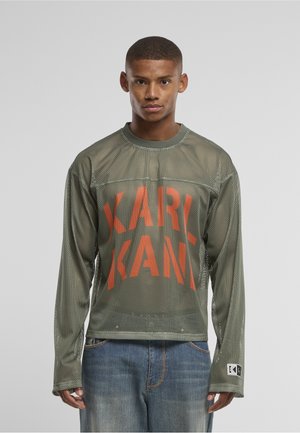 Ung mand iført en grøn netlangærmet skjorte med stor orange "KARL KANI" tekst og blå jeans, stående mod en ensfarvet baggrund.