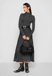 Abito maxi nero plissettato con scintillio metallico, maniche lunghe e spalle scoperte, abbinato a una cintura nera e una borsa strutturata.