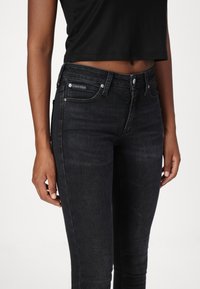 Jeans skinny de mezclilla negra con una cintura de altura media, que presentan una etiqueta de marca, bolsillos frontales y detalles sutiles de decoloración.