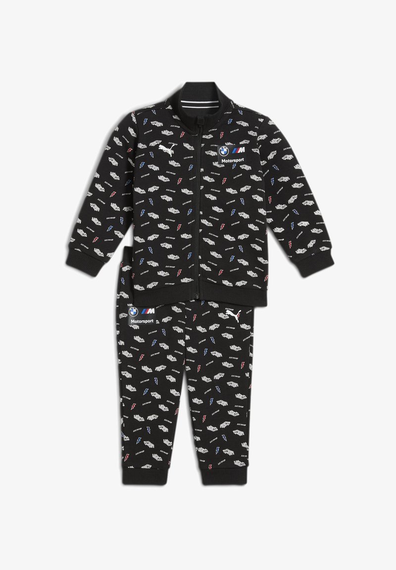 Juodas treniruočių kostiumas, kuriame yra užtrauktuku užsegamas švarkas ir kelnės. Audinys yra spausdintas su mažais baltais automobiliais ir žaibais mėlynos ir raudonos spalvų.