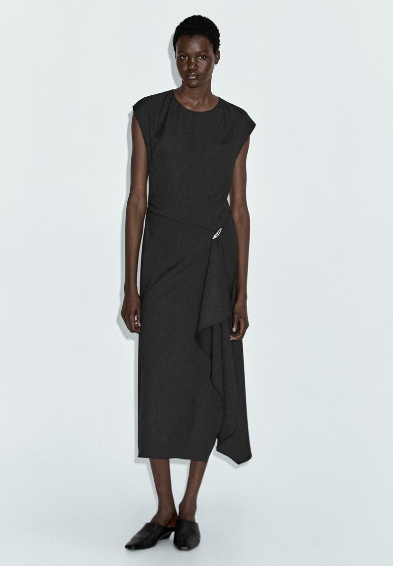 Schwarzes, knielanges Kleid mit kurzen Ärmeln, drapiertem Design und seitlichem Twist-Akzent; strukturierter Stoff mit glatter Oberfläche.