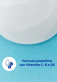 Contenitore circolare bianco con una superficie liscia, posizionato su uno sfondo turchese. Il testo sottolinea una formula protettiva con vitamine C, E e oli.