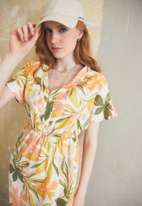 Virágmintás, könnyű anyagból készült ruha, narancssárga, rózsaszín és zöld trópusi mintákkal. V-nyakú dekoltázs és gumis derék a laza illeszkedés érdekében.