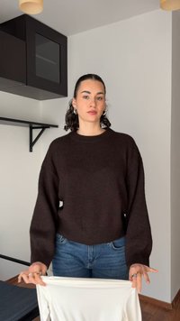 Dark brown knitted sweater with a rounded neckline and long, wide sleeves, paired with blue denim jeans, held over white fabric.