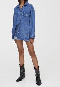 Persona que lleva una chaqueta de denim azul de gran tamaño, pantalones cortos de denim a juego y botas vaqueras negras con costuras decorativas, de pie sobre un fondo blanco.