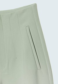 Pantalon sur mesure en tissu doux vert clair, présentant une texture lisse, un devant plat et un détail discret de poche latérale.