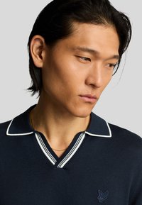 Polo shirt a maglia blu navy con colletto a righe bianche, caratterizzato da un design a scollo a V e un piccolo logo ricamato sul petto.