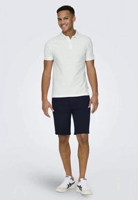 Hvit polo skjorte for menn med liten krage, kombinert med marineblå shorts og hvite sneakers med svarte detaljer. Glatt stoff, avslappet passform.