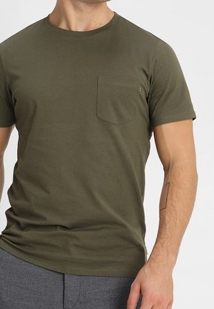 Homme portant un t-shirt vert olive à manches courtes avec une poche poitrine et un pantalon gris, montrant un simple tatouage linéaire sur son avant-bras.