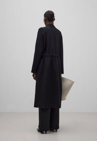 Musta pitkä takki, jossa on vyötäröltä muotoiltu leikkaus, pitkät hihat ja sileä pinta. Yhdistetty leveälahkeisiin housuihin ja vaaleanväriseen laukkuun.