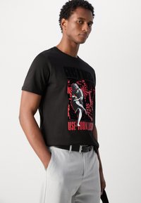 Black cotton t-shirt featuring a central graphic in red and white, with bold text, paired with light grey trousers and a black belt.