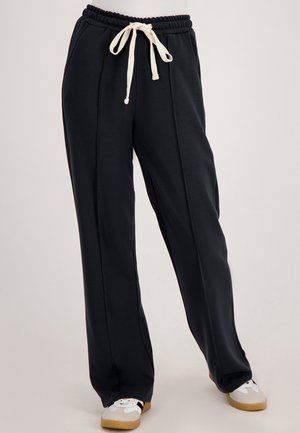 Zwarte wijduitlopende broek met voornaad, elastische tailleband en crèmekleurige trekkoord, gecombineerd met witte en beige sneakers op een neutrale achtergrond.