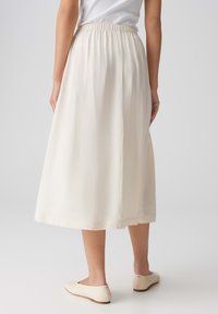 Jupe de couleur crème, de longueur mi-mollet, avec une taille élastique. Texture lisse, design fluide et un éclat subtil, associée à des ballerines de couleur claire.