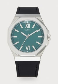 Teal dials klokke fra Guess med sølvfarget oktagonal hus, svart gummireim og hvite timemarkører ved VI og XII. Har minimalistisk design.