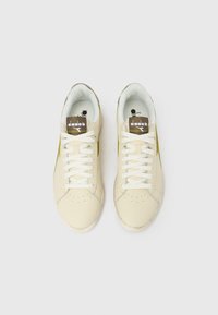 Ζευγάρι κρεμ χρωματιστά αθλητικά παπούτσια Diadora με λευκά κορδόνια και καφέ ετικέτες λογότυπου, εμφανιζόμενα από πάνω σε λευκό φόντο.