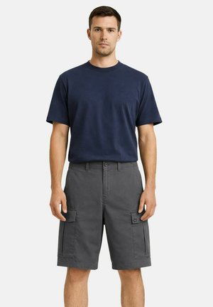 Mann trägt ein marineblaues T-Shirt und graue Cargo-Shorts, steht vor einem einfarbigen hellen Hintergrund.