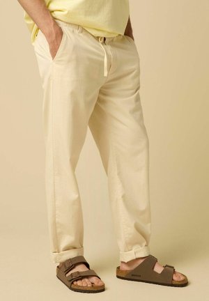 Mann trägt hellbeige Hose mit Händen in den Taschen und braune Birkenstock-Sandalen vor beigem Hintergrund in passender Farbe.
