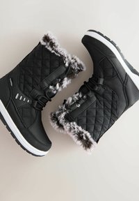 Czarne pikowane zimowe botki z futrzanym wykończeniem, sznurówki z przodu, etykieta wodoodporna oraz grube białe akcentowane podeszwy na jasnoszarym tle.