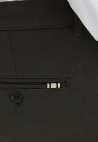 Dunkelgrüne Hosen mit strukturiertem Finish, die über eine kleine Vordertasche mit gestreiftem Akzent und einem einzelnen schwarzen Knopfverschluss verfügen.