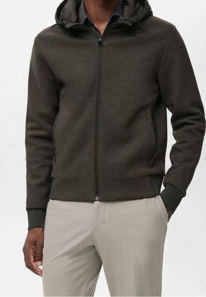 Homme portant un sweat à capuche zippé vert olive foncé avec des poignets côtelés et un pantalon beige, une main dans la poche et fond neutre.