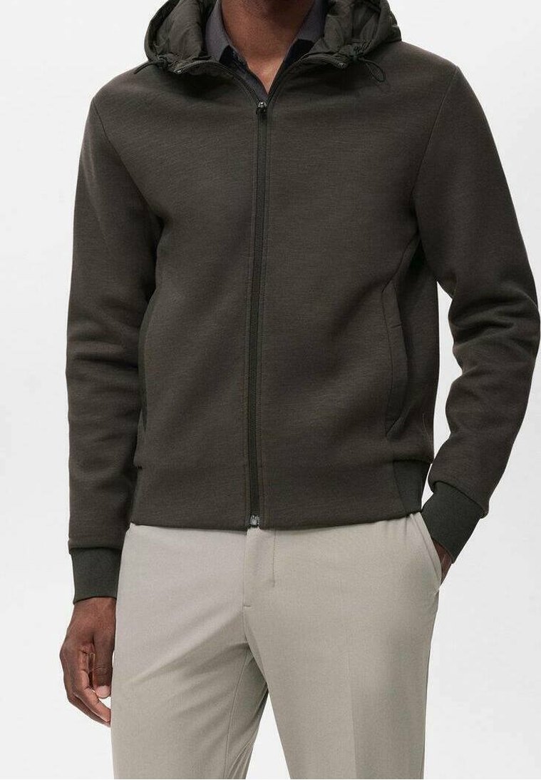 Homme portant un sweat à capuche zippé vert olive foncé avec des poignets côtelés et un pantalon beige, une main dans la poche et fond neutre.
