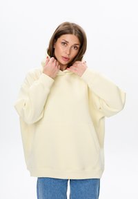Felpa con cappuccio oversize di colore giallo pallido, con tasca frontale e polsini a costine. Il tessuto sembra morbido con una texture liscia.