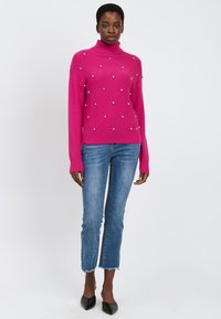 Fuchsia gestrickter Pullover mit hochgeschlossenem Kragen, verziert mit diagonalem Rautenmuster und metallischen Akzenten. Kombiniert mit blauen Jeans und schwarzen Pumps.