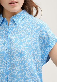 Blusa a maniche corte realizzata in tessuto azzurro chiaro con motivo floreale. Presenta un colletto e una chiusura con bottoni. Texture liscia.